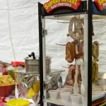A concession stand setup featuring a SuperPretzel soft pretzel warmer with pretzels inside, tongs, small cups, a pot, chips, yellow plates, and a red-and-white tablecloth.