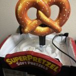 A plastic display featuring a large, realistic pretzel sits on top of a SuperPretzel Soft Pretzels warming machine, with cords visible and a beige wall in the background.