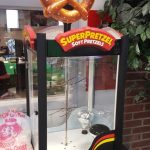 A SuperPretzel display case with a large pretzel sign on top stands empty on a table. The case is labeled "Soft Pretzels" and sits next to a pink plastic bag and a potted plant.
