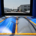View from inside a blue and gray inflatable bounce house, looking out through the netted entrance toward industrial buildings and a clear sky in the background.