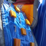 Close-up of a colorful inflatable climbing wall with blue and orange sections, yellow footholds, and silver handles, likely part of a bouncy house or inflatable play structure for children.