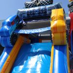 An inflatable blue and yellow slide designed to look like a monster truck, with large wheels and a racing-themed decoration at the top, set outdoors on a sunny day.