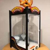 A glass display case for SuperPretzel soft pretzels sits on a table, featuring a large plastic pretzel on top and empty shelves inside. The case has black trim and a colorful stripe along the bottom.