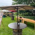 A round table made from a wooden barrel with a large beige umbrella stands on green grass. Wooden benches and more barrel tables are set up outdoors under trees, suggesting an outdoor event or picnic.