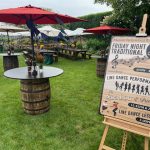 Outdoor event with barrel tables and picnic benches under red umbrellas. A sign advertises live music and line dance performances by Friday Night Traditional, Sara Lout & Friends, and a line dance lesson.