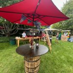 A wooden barrel table with flowers and a lantern sits under a large red umbrella on a grassy lawn. In the background, people stand near food stalls and trees.