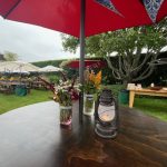 A round wooden table with a lantern and jars of flowers sits under a red umbrella in an outdoor garden with picnic tables, more umbrellas, and lush greenery in the background.
