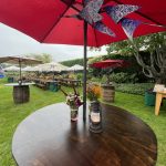 A round wooden table with a lantern and flower vase sits under a large red umbrella decorated with bunting. More tables with umbrellas are set up on green grass in a garden setting.