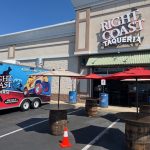 Outdoor scene of Right Coast Taqueria restaurant with a colorful food truck and three tables made from barrels with umbrellas. Orange traffic cones mark the area in a parking lot on a sunny day.