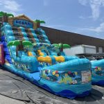 Large inflatable water slide features a beach theme, vibrant palm trees, and a visible "Beach Party" sign.