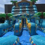 An inflatable water slide with a "Beach Party" sign features palm trees and tropical designs under the sun.