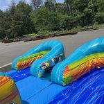 A colorful inflatable water slide sits in a parking lot, surrounded by trees and metal beams in the background.