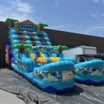 Large inflatable water slide with a “BEACH” sign and tropical decorations stands outdoors on pavement.